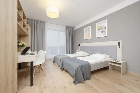 A cozy bedroom featuring a single bed with a neutral-toned duvet, a wooden desk, and a large window allowing natural light to brighten the space.

