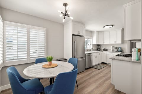 Bright kitchen and dining area with white shaker cabinets, stainless steel appliances, and marble countertops. Modern chandelier hangs over a round marble table with blue chairs. Large windows with shutters fill the space with natural light.