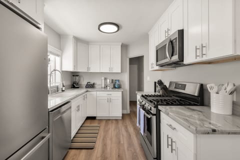 Bright kitchen and dining area with white shaker cabinets, stainless steel appliances, and marble countertops. Modern chandelier hangs over a round marble table with blue chairs. Large windows with shutters fill the space with natural light.