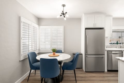 The dining area features a round marble table with blue chairs near large windows. Stainless steel appliances, white shaker cabinets, and wood-look flooring complete the space.