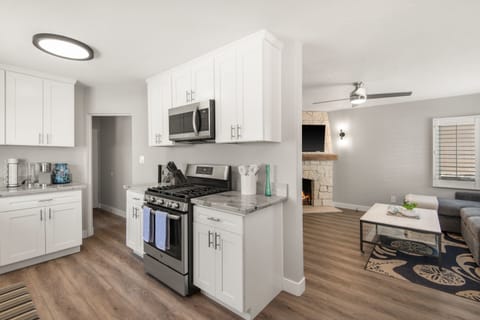 Bright kitchen and dining area with white shaker cabinets, stainless steel appliances, and marble countertops. Modern chandelier hangs over a round marble table with blue chairs. Large windows with shutters fill the space with natural light.