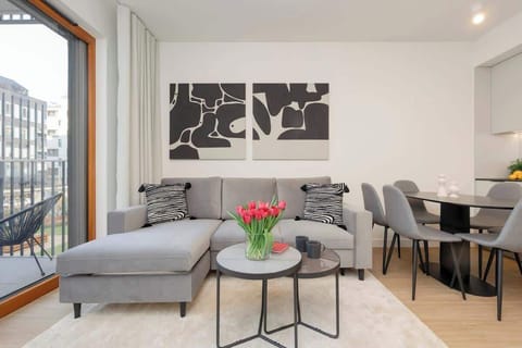 A stylish living room with a gray sofa, a glass coffee table, and modern black-and-white wall art. A floor-to-ceiling window allows natural light to brighten the space.

