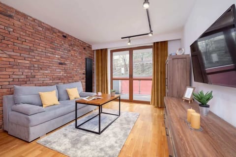 A stylish living room with an exposed brick wall, a gray sofa, a modern coffee table, and large windows with floor-length curtains.

