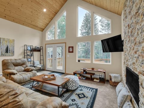 Sun-drenched second-level living room featuring vaulted pine ceilings, floor-to-ceiling forest views, and a cozy stone fireplace