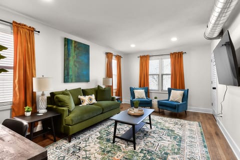 Sink into the plush green sofa and unwind in your stylish living room, where vibrant orange curtains frame windows and cozy blue chairs invite relaxation.