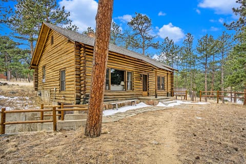 A rustic log cabin surrounded by pine trees on a clear day, with a visible dirt path and wooden fencing.