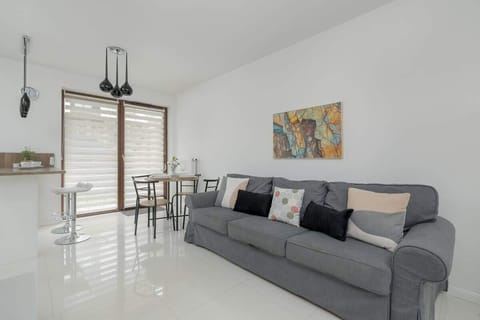 A bright living room with a gray sofa, a small round table, and large glass doors leading to the terrace. The space is minimalistic and modern.

