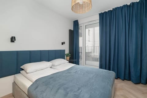A modern bedroom area with a large bed featuring blue bedspreads and pillows. The decor is simple, with dark blue curtains and light walls, creating a calming ambiance.