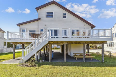 A charming coastal-style home elevated on stilts features a spacious wrap-around deck and covered ground-level area with picnic seating, nestled in a quiet residential neighborhood.