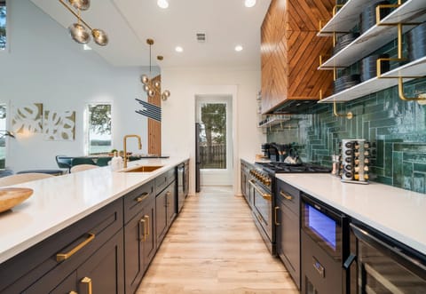 A Stylish Kitchen Space Featuring Beautiful Cabinetry, Elegant Gold Accents, White Countertops, And Floating Wooden Shelves, With Natural Light Streaming In Through A Door At The End Of A Galley Setup.