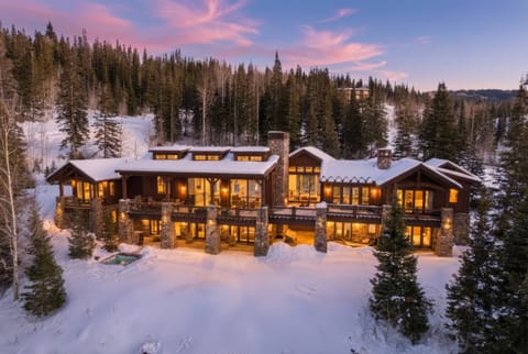 Twilight exterior view of luxury mountain home:
Snow-covered luxury cabin at dusk with glowing interior lights, surrounded by pines and mountains under a purple sk