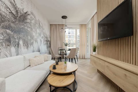 A modern living room with light-toned furniture and wood-panel walls. Large windows let in natural light, making the space bright and inviting.