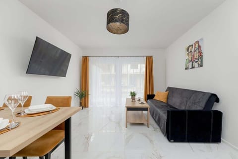 A bright, modern space featuring a black sofa, a small wooden coffee table, and a large wall-mounted TV. The room has a minimalist design with light-colored floors and walls, creating a spacious atmosphere.