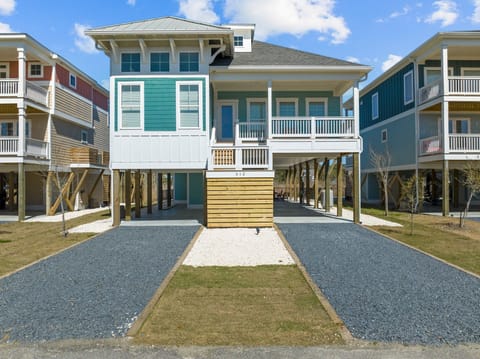 Teal raised beach house with white railings, covered porch, gravel parking, wood privacy panel, and coastal curb appeal for an Oak Island, NC vacation rental.