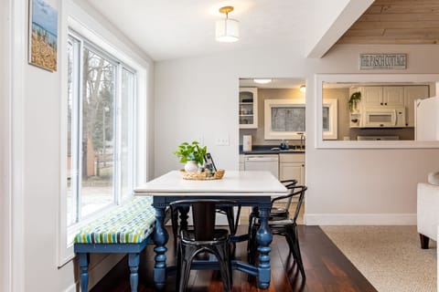 Experience modern elegance and traditional charm in our dining area. A white table, black chairs, and a bench with a green striped cushion are bathed in natural light from large windows. A quaint sign reading 'THE BEACH COTTAGE' adds a charming touch.