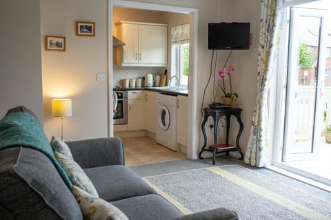 Lounge area leading to open kitchen. 2 seater sofa with cushions. TV on wall, small table