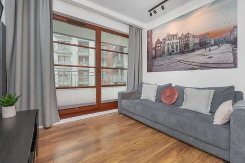 A bright, modern living room with a grey sofa, wooden flooring, large windows, and a wall-mounted TV.