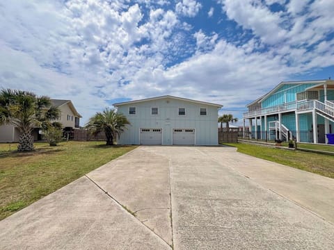 A coastal-style property featuring a spacious two-car garage and convenient parking, nestled in a charming neighborhood with palm trees and neighboring beach homes under a bright, cloud-swept sky.