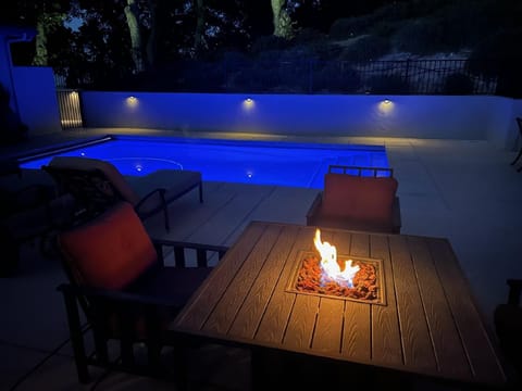 As evening falls, the poolside terrace transforms into an enchanting escape, featuring a glowing fire pit and a pool illuminated with a deep blue hue. Subtle wall lighting enhances the ambience, creating a perfect setting for great nighttime relaxation.
