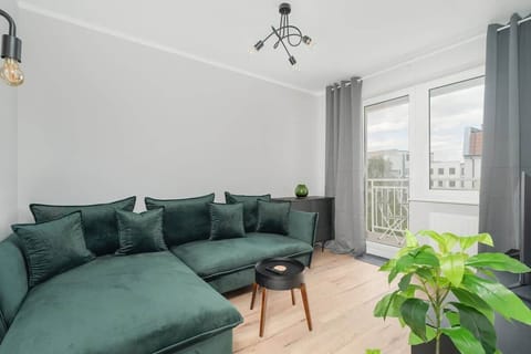 A bright living room with a large green sectional sofa, a small coffee table, and potted plants by the window.