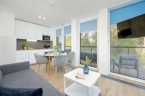 The living room is connected to the kitchenette. In between stands a table with a round top. The room has large windows and access to the balcony. 