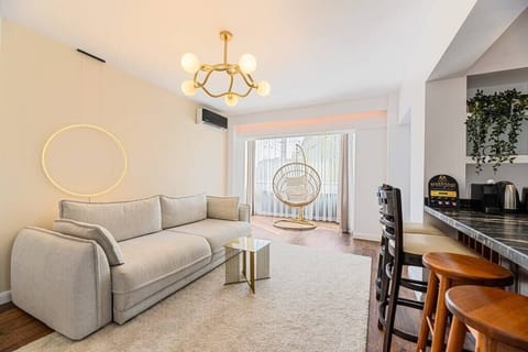 Stylish living room featuring a huge extendable gray sofa, illuminating lamp, cozy chair with pillow on it, AC, high chairs and table for dining in the open space kitchen 