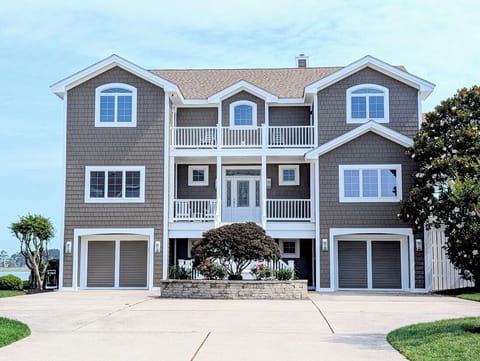 33 Inlet View North Bethany Beach Exterior - A front view of a multi-story residence with gray shingle siding, a large concrete driveway, two garages, and an upper-level balcony with white railings.