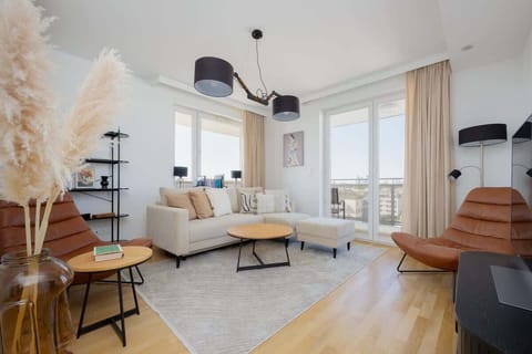 A bright living area with large windows, beige furnishings, and an open-plan kitchen and dining zone.

