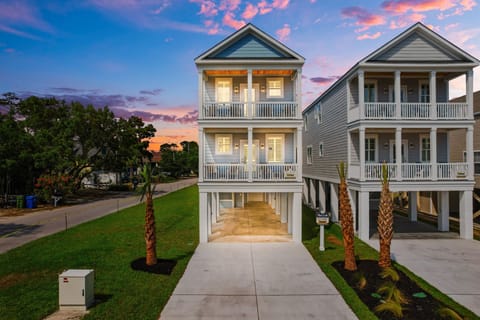 Stunning coastal rental featuring elegant architecture with wrap-around porches and palm-lined entrance under brilliant sunset skies.