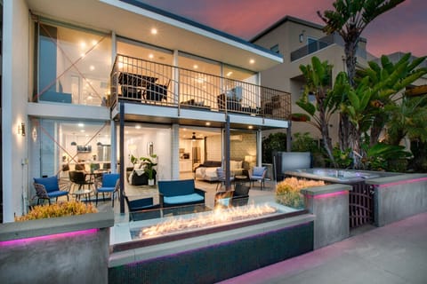 A luxurious two-story beachfront home illuminated at sunset, featuring expansive glass windows, multiple outdoor seating areas, and a shared hot tub. This stunning property offers a perfect blend of relaxation and sophistication with breathtaking views of