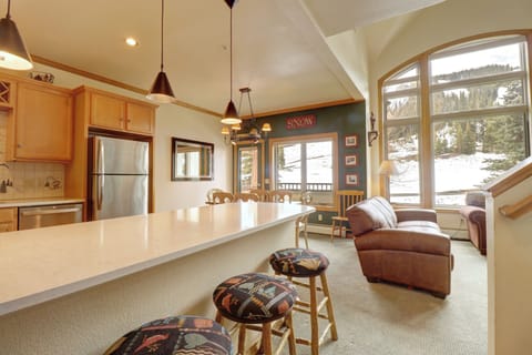 A cozy kitchen area with a long countertop and bar stools, next to a living room featuring a leather couch and large windows overlooking a snowy landscape.
