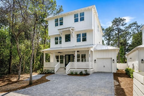 Modern three-story coastal home features wrap-around porch and garage, nestled among mature trees in peaceful residential setting.