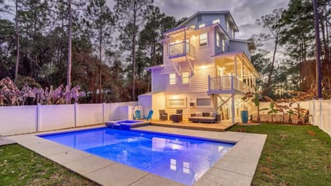 Modern multi-level beach house with illuminated pool area, surrounded by pine trees and featuring multiple decks and balconies.