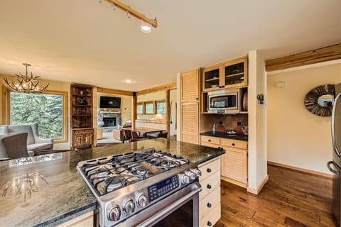 A modern kitchen with a stainless steel gas stove, granite countertops, wooden cabinetry, and an open layout leading to a living area with large windows and forest views.