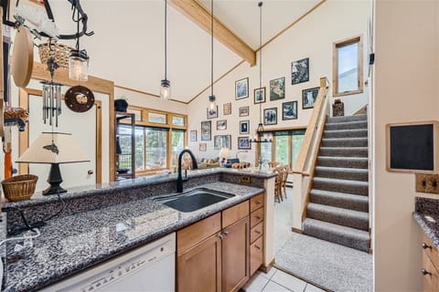 A kitchen with granite countertops, wooden cabinets, and a sink. Stairs lead upstairs on the right, and a wall decorated with framed pictures is visible in the background.