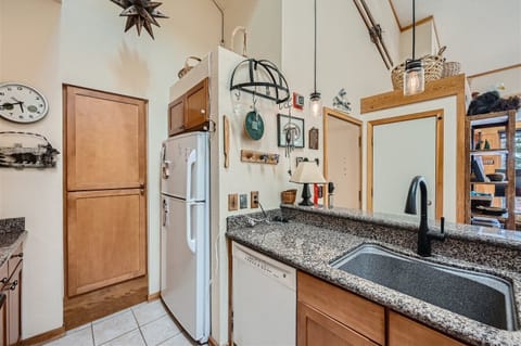A compact kitchen featuring a granite countertop with a sink and a black faucet, white refrigerator, wooden cabinets, dishwasher, and various hanging utensils and decor on the wall.