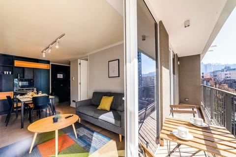 Modern studio living room with a colorful geometric rug, full-height glass doors, and a furnished balcony overlooking the city and surrounding hillside.