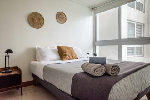 Private bedroom with crisp layered bedding, rolled towels, and a large window bringing natural daylight into the studio.