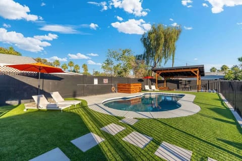 Sunlit backyard with a curved pool, loungers, and bright umbrellas—start with a refreshing dip, then stretch out on the green turf with the blue sky overhead.