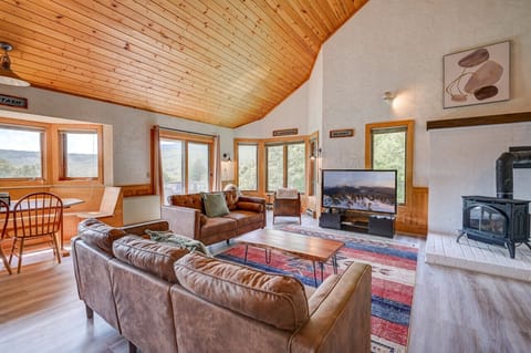 Spacious open-concept living area with vaulted wood ceilings, large TV, gas wood stove, AC, and cozy seating. Enjoy natural light, forest views, and rustic-modern charm—perfect for relaxing