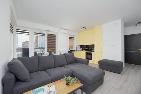 A contemporary living room with a spacious gray sectional sofa, light flooring, and a pop of yellow in the kitchen cabinetry.
