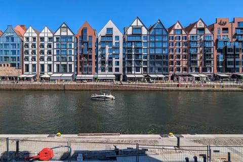 A waterfront view of colorful Dutch-style buildings reflected in the river, with boats docked in the foreground. The blue sky enhances the charm of the urban landscape.
