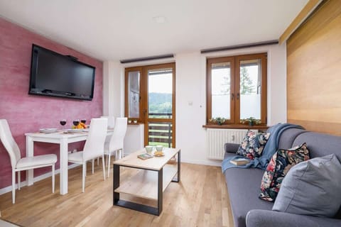 A well-lit lounge with wooden accents, a wall-mounted TV, and a compact dining table near a window, creating a warm and inviting atmosphere.