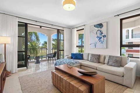This spacious, well-lit living room has a modern vibe with a comfy sectional sofa, coffee table, and large flat-screen TV. It's decorated in neutral colors with blue accents and has a sliding glass door that opens to a balcony with pool and greenery 