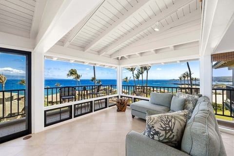 Spacious covered lanai featuring cushioned lounge seating and a dining table, overlooking lush tropical landscaping and expansive blue ocean views beyond the balcony railing.