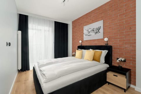 A modern bedroom with a black bed frame, light wooden furniture, and a large window with dark curtains. Exposed brick adds warmth and texture to the design.