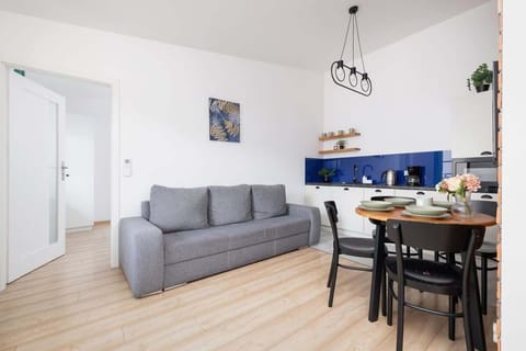 A cozy living room space with a gray sofa, small dining table, and minimalist decor. Natural light fills the room through a large window.