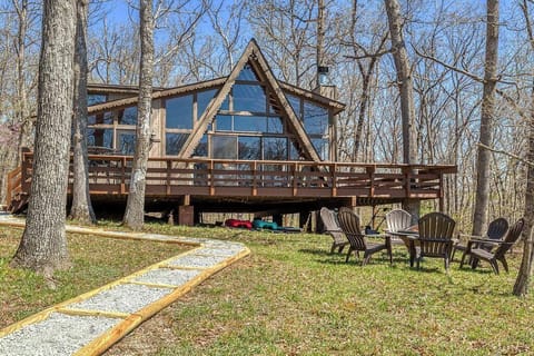 Full exterior view of the glass-fronted A-frame cabin with a broad wood deck, gravel path and circular outdoor seating among mature trees.