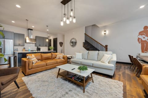 The warm living room features a leather couch, open kitchen, and a lush plant that sets a welcoming tone.