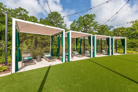 There are several modern cabana-like structures or pergolas. They have white frames, light-colored slatted roofs, and some feature dark green curtains that can be drawn for privacy or shade.